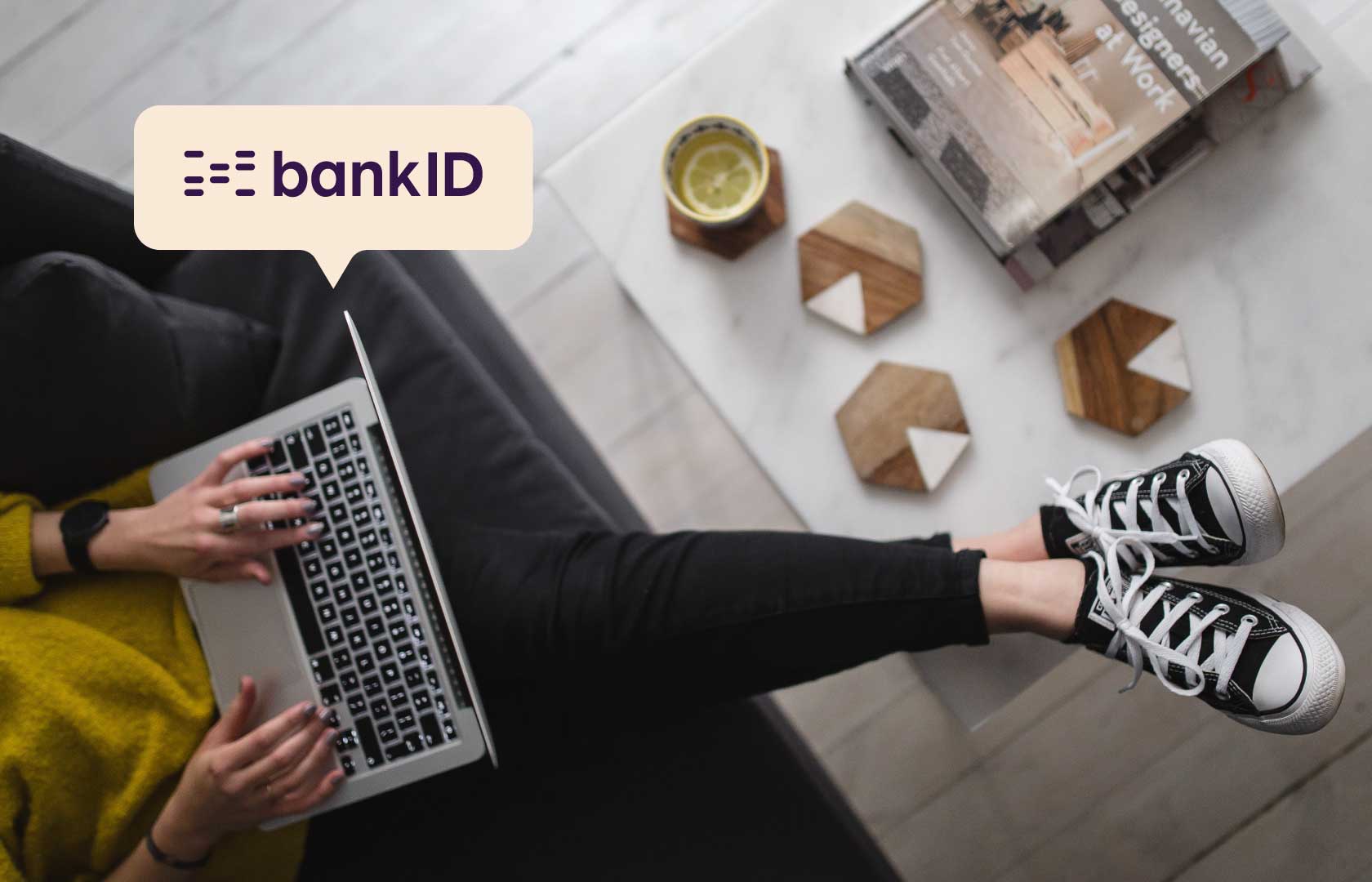 Foto fulgeperspektiv, kvinne i sofa med Mac på fanget, hviler beina på et kaffebord, grafikk snakkeboble med logo av BankID